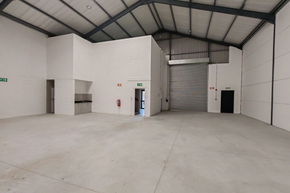 Spacious industrial warehouse unit featuring high ceilings with exposed steel trusses, concrete flooring, a roller shutter door, and a small integrated kitchenette area.