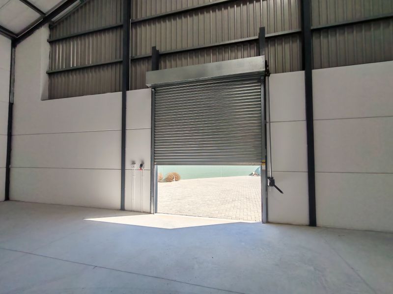 Spacious industrial utility area featuring high-clearance ceilings, concrete flooring, and a large roller shutter door providing direct access to the exterior paved yard.