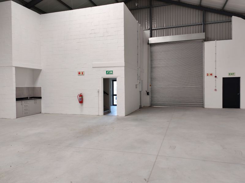Spacious industrial warehouse unit featuring high ceilings, concrete flooring, a large roller shutter door, and a functional kitchenette area.