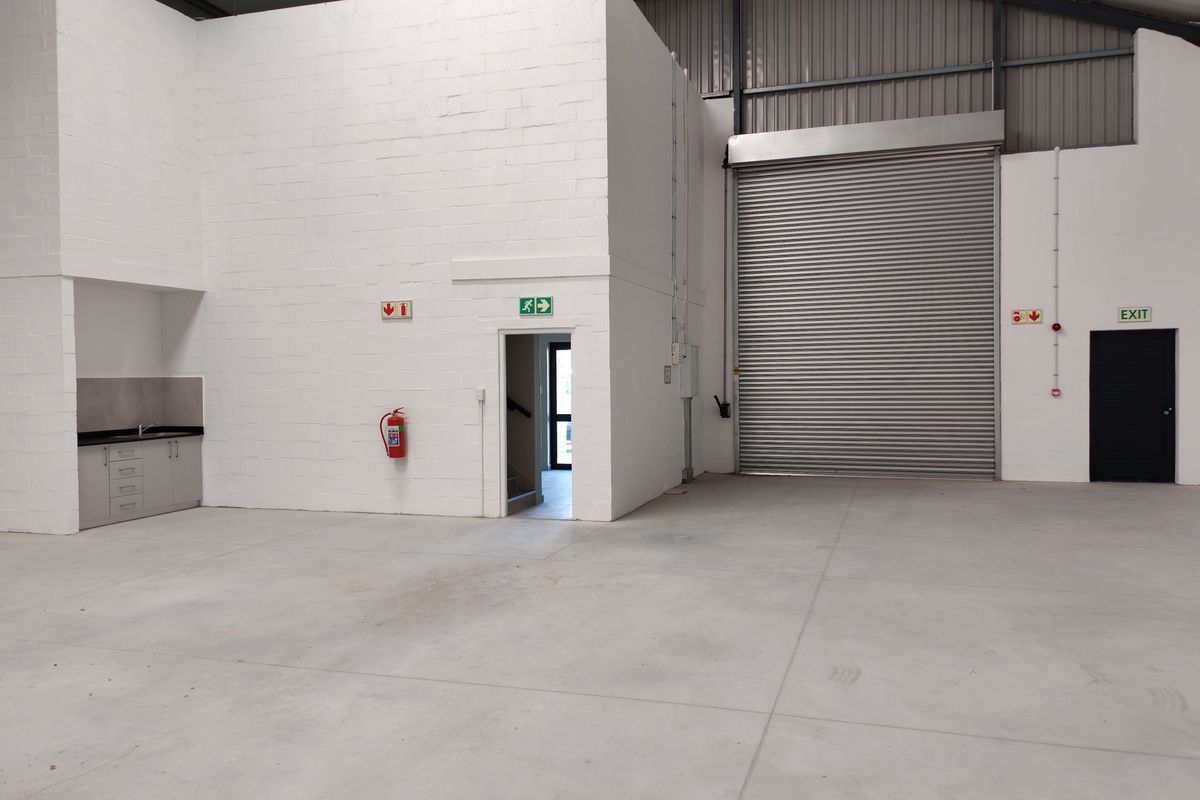 Spacious industrial warehouse unit featuring high ceilings, concrete flooring, a large roller shutter door, and a functional kitchenette area.