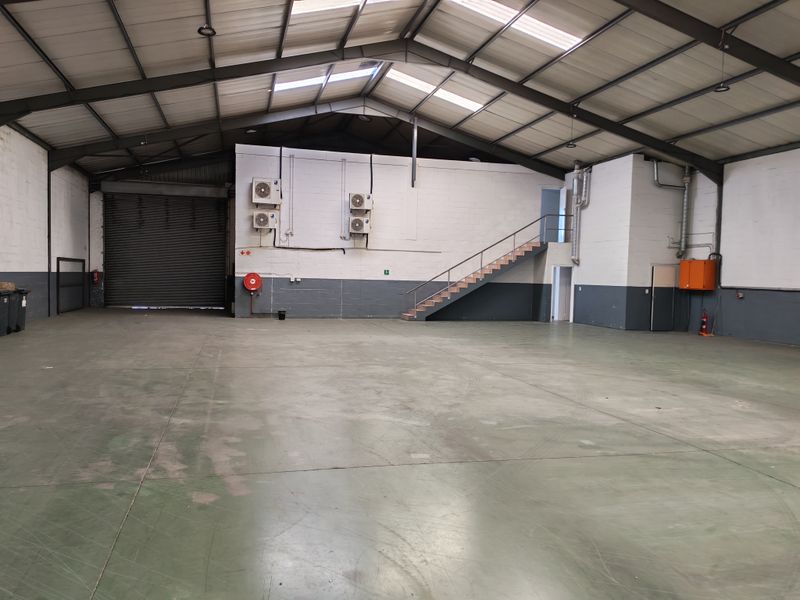 Spacious industrial warehouse unit featuring high ceilings with skylights, a roller shutter door, internal staircase, and wall-mounted air conditioning units.