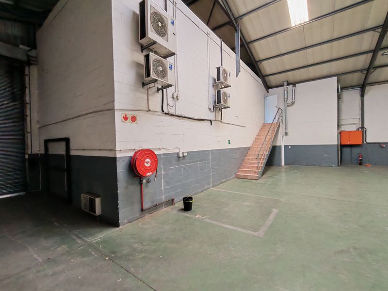 Spacious industrial warehouse unit featuring high ceilings with exposed steel trusses, concrete flooring, fire safety equipment, and wall-mounted air conditioning units.