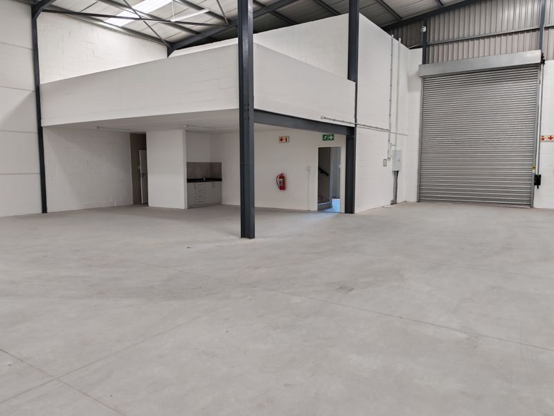 Spacious industrial warehouse unit featuring a large roller shutter door, mezzanine level, concrete flooring, and a functional kitchenette area.