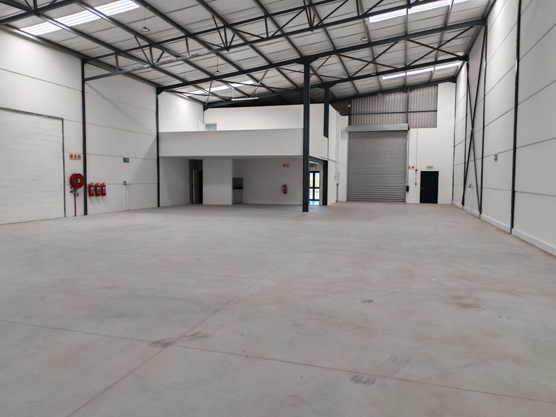 Spacious industrial warehouse unit featuring high ceilings with exposed steel trusses, a mezzanine level, concrete flooring, and a large roller shutter door.