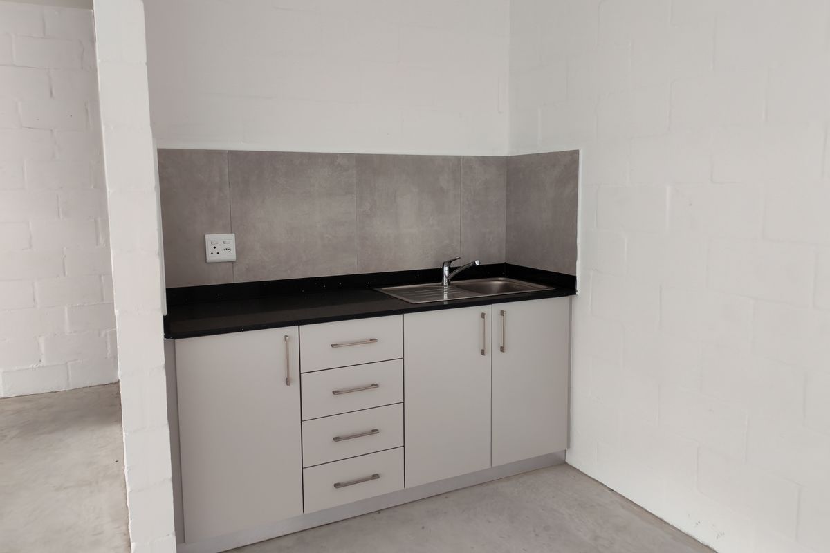 A clean, compact kitchenette featuring modern grey cabinetry, a dark stone countertop, stainless steel sink, and a neutral tiled backsplash in an industrial space.