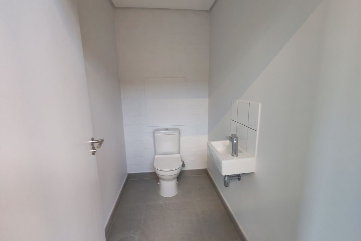 A clean and functional industrial-style restroom featuring a modern toilet, wall-mounted basin with chrome tap, and neutral tiled flooring.