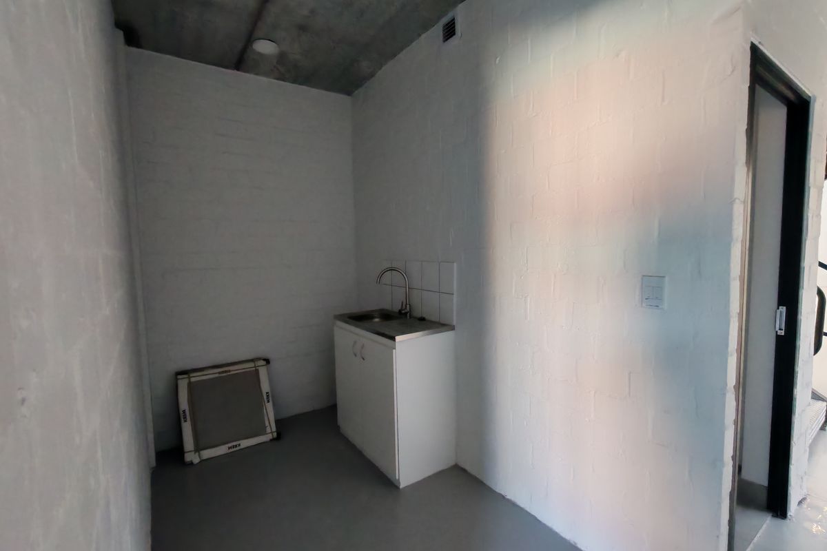 A compact industrial-style kitchenette featuring a stainless steel sink, built-in cabinetry, tiled splashback, and exposed concrete ceiling in a minimalist white brick space.