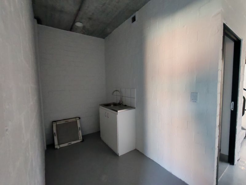 A compact industrial-style kitchenette featuring a stainless steel sink, built-in cabinetry, tiled splashback, and exposed concrete ceiling in a minimalist white brick space.