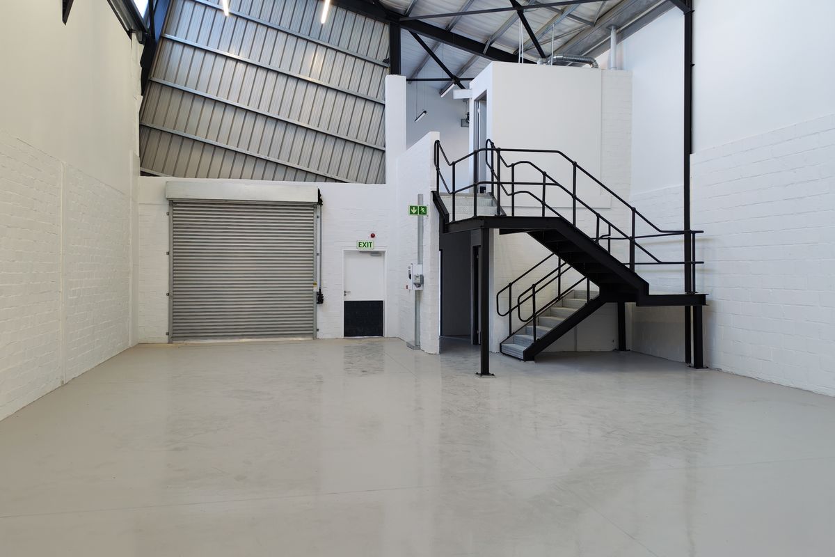 Spacious industrial warehouse unit featuring high ceilings with exposed steel trusses, a roller shutter door, and a mezzanine level accessed by a metal staircase.