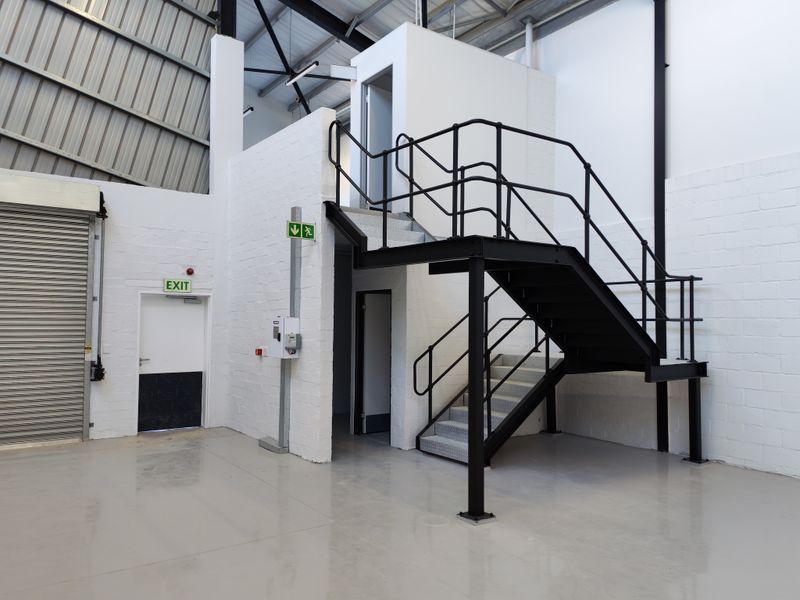 Industrial warehouse interior featuring high ceilings, epoxy flooring, a roller shutter door, and a metal staircase leading to an elevated mezzanine level.