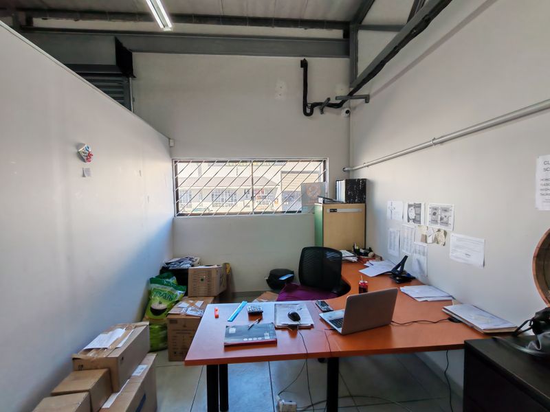 A functional industrial office space featuring exposed steel roof beams, security-barred windows, and basic utility finishes, suitable for administrative or operational use.