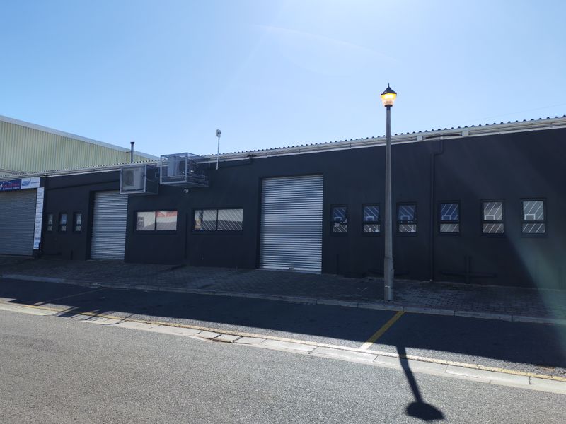 Industrial property exterior featuring dark painted facade, multiple roller shutter doors, and external air conditioning units with paved street-level access.