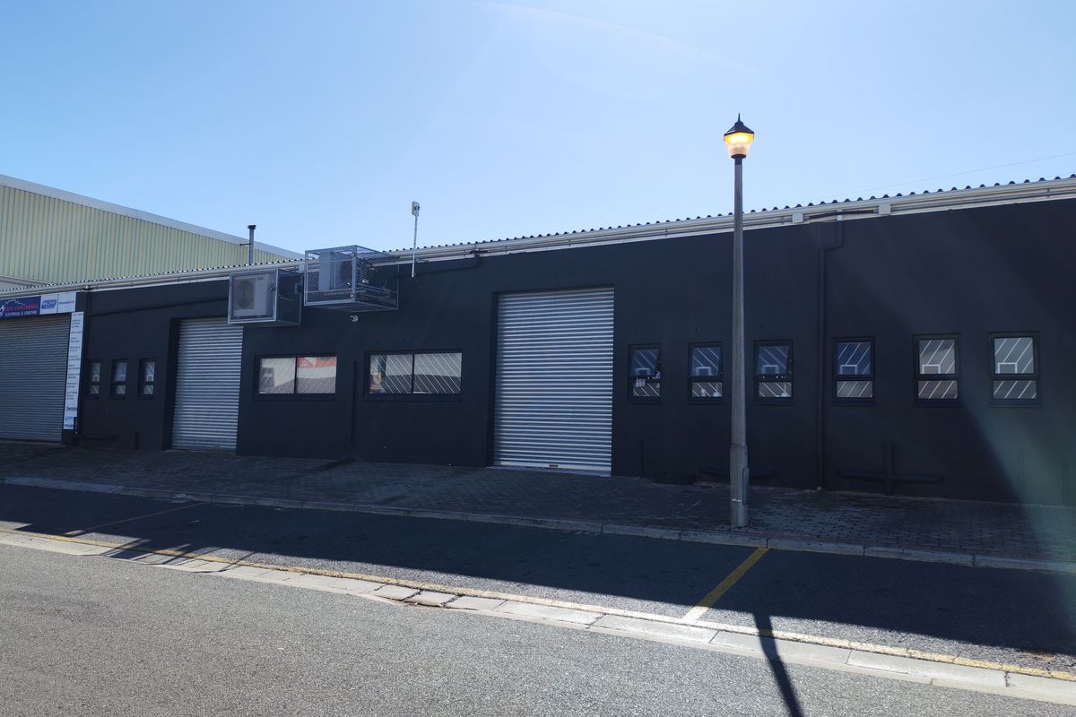 Industrial property exterior featuring dark painted facade, multiple roller shutter doors, and external air conditioning units with paved street-level access.