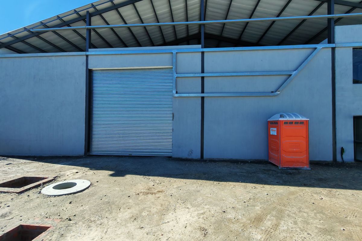 Exterior view of an industrial warehouse featuring a large roller shutter door, steel structural framing, and an unfinished surrounding ground area.