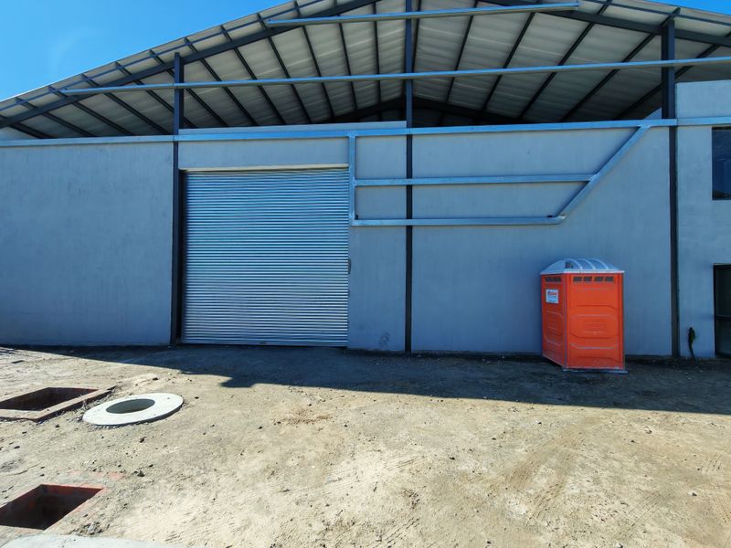 Exterior view of an industrial warehouse featuring a large roller shutter door, steel structural framing, and an unfinished surrounding ground area.