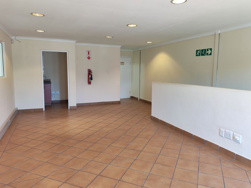 An open-plan office space featuring ceramic tiled flooring, recessed lighting, and essential safety fixtures including a fire extinguisher and emergency exit signage.