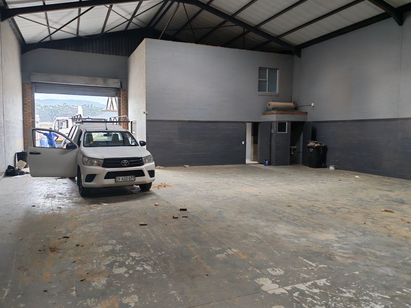 Spacious industrial warehouse unit featuring high ceilings with exposed steel trusses, concrete flooring, and a large roller shutter door for vehicle access.