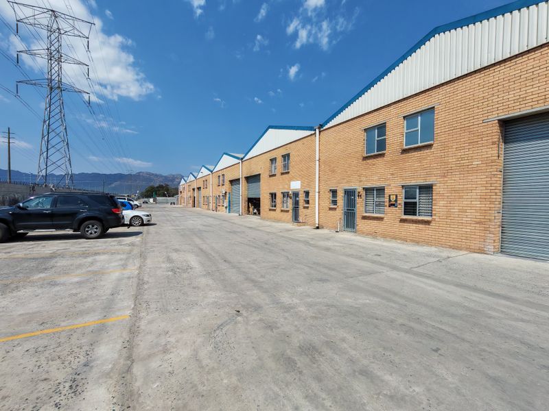 Exterior view of an industrial warehouse complex featuring face brick construction, roller shutter doors, and a paved concrete driveway for vehicle access.