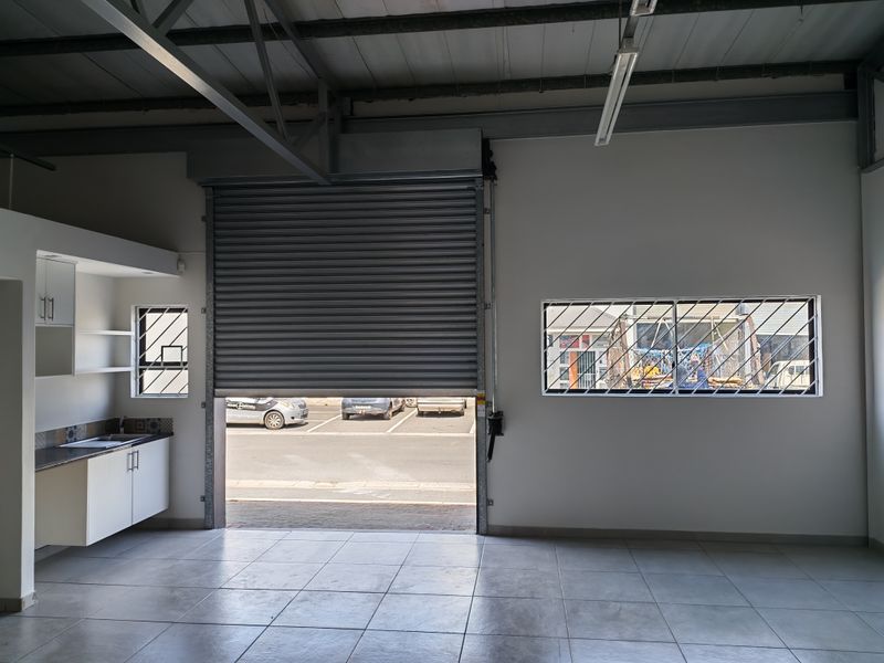 Industrial utility space featuring a large roller shutter door, tiled flooring, and a compact built-in kitchenette with security bars on windows.
