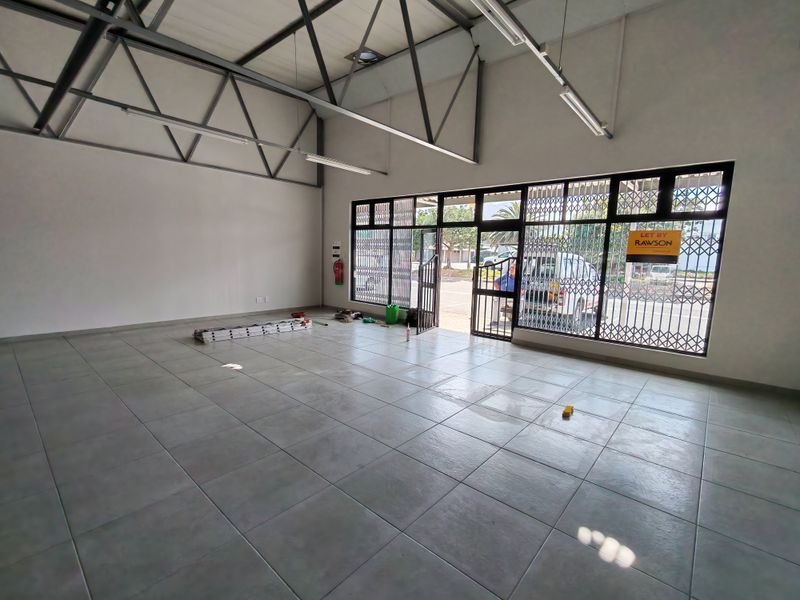 Spacious open-plan industrial unit featuring high exposed steel trusses, large glass storefront windows with security gates, and durable tiled flooring.