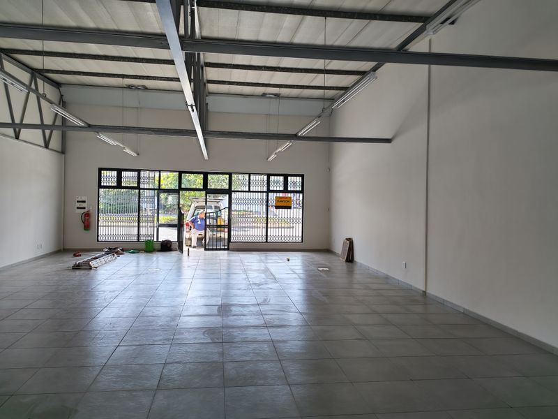 Spacious open-plan industrial unit featuring tiled flooring, high exposed steel roof trusses, and a large glass storefront with security gate access.