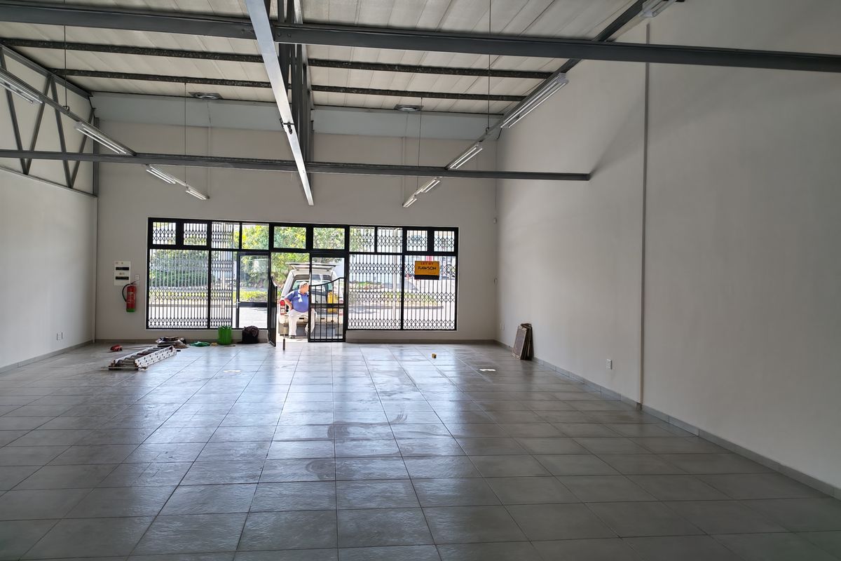 Spacious open-plan industrial unit featuring tiled flooring, high exposed steel roof trusses, and a large glass storefront with security gate access.