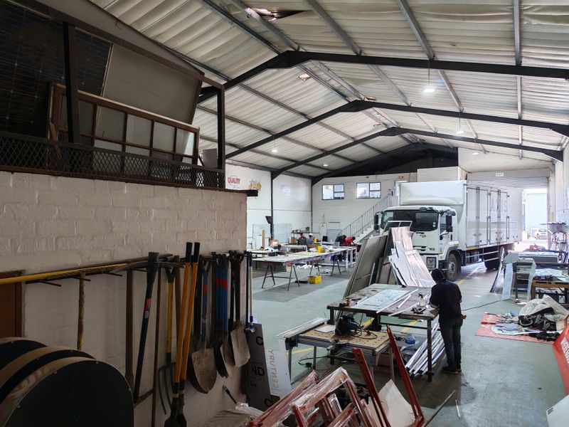 Spacious industrial warehouse featuring high ceilings with exposed steel trusses, concrete flooring, a mezzanine level, and a large roller shutter door for vehicle access.