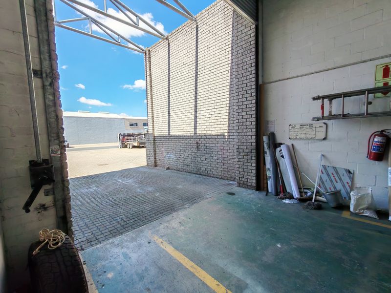 Industrial garage space featuring high ceilings with exposed steel trusses, painted brick walls, and a large roller shutter door opening to a paved driveway.