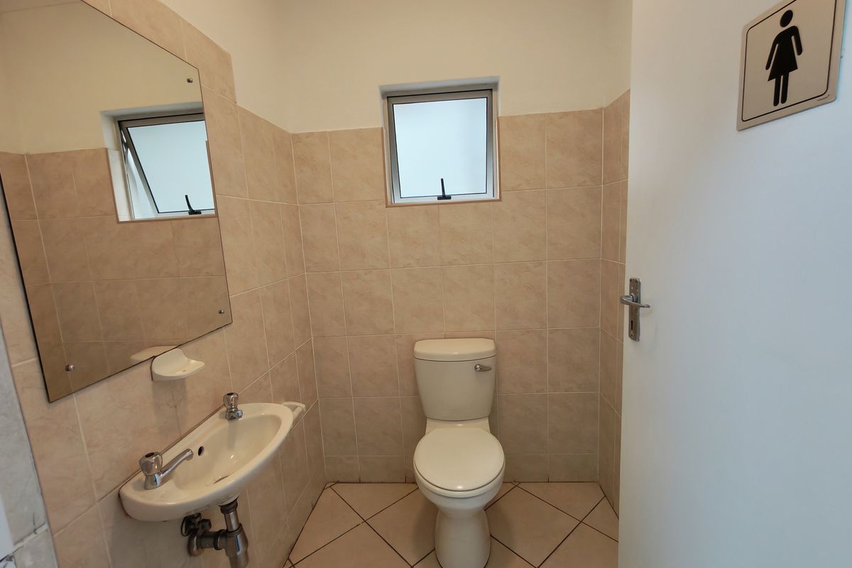 A functional industrial restroom featuring ceramic wall tiles, a wall-mounted basin, a standard toilet, and a small window for ventilation.
