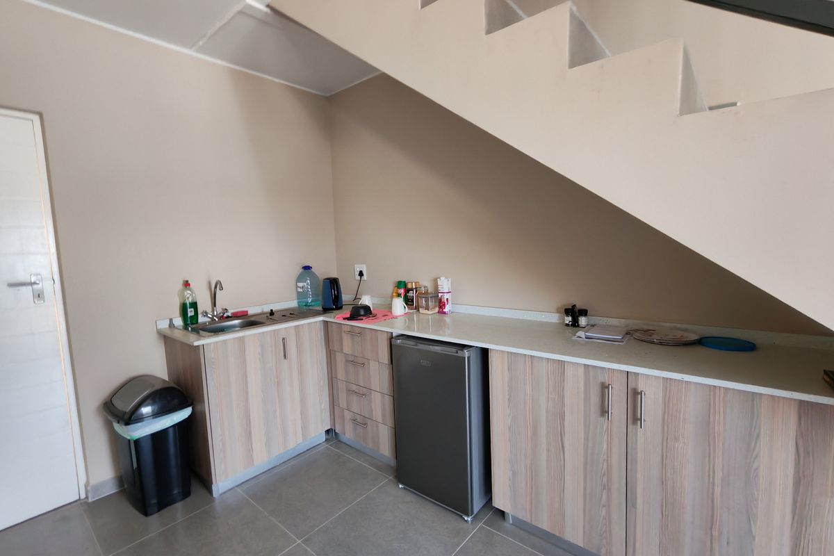 A compact kitchenette area featuring light wood-grain cabinetry, a stainless steel sink, and a stone-look countertop, conveniently located beneath a staircase.