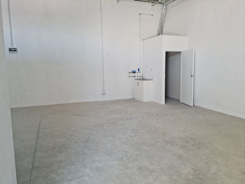 An open-plan industrial utility space featuring concrete flooring, exposed ducting, and a small built-in sink cabinet with plumbing connections.