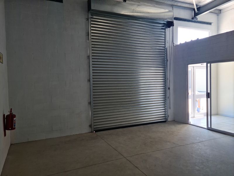 Industrial garage space featuring a large roller shutter door, polished concrete flooring, exposed steel structural beams, and a sliding glass door entrance.