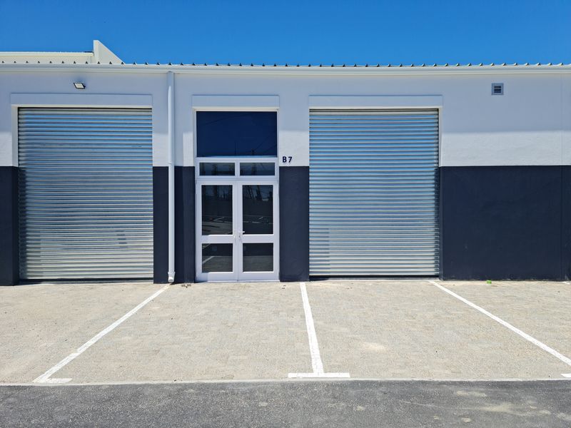 Modern industrial unit facade featuring two secure roller shutter doors, a central glass entrance, and dedicated asphalt parking bays.