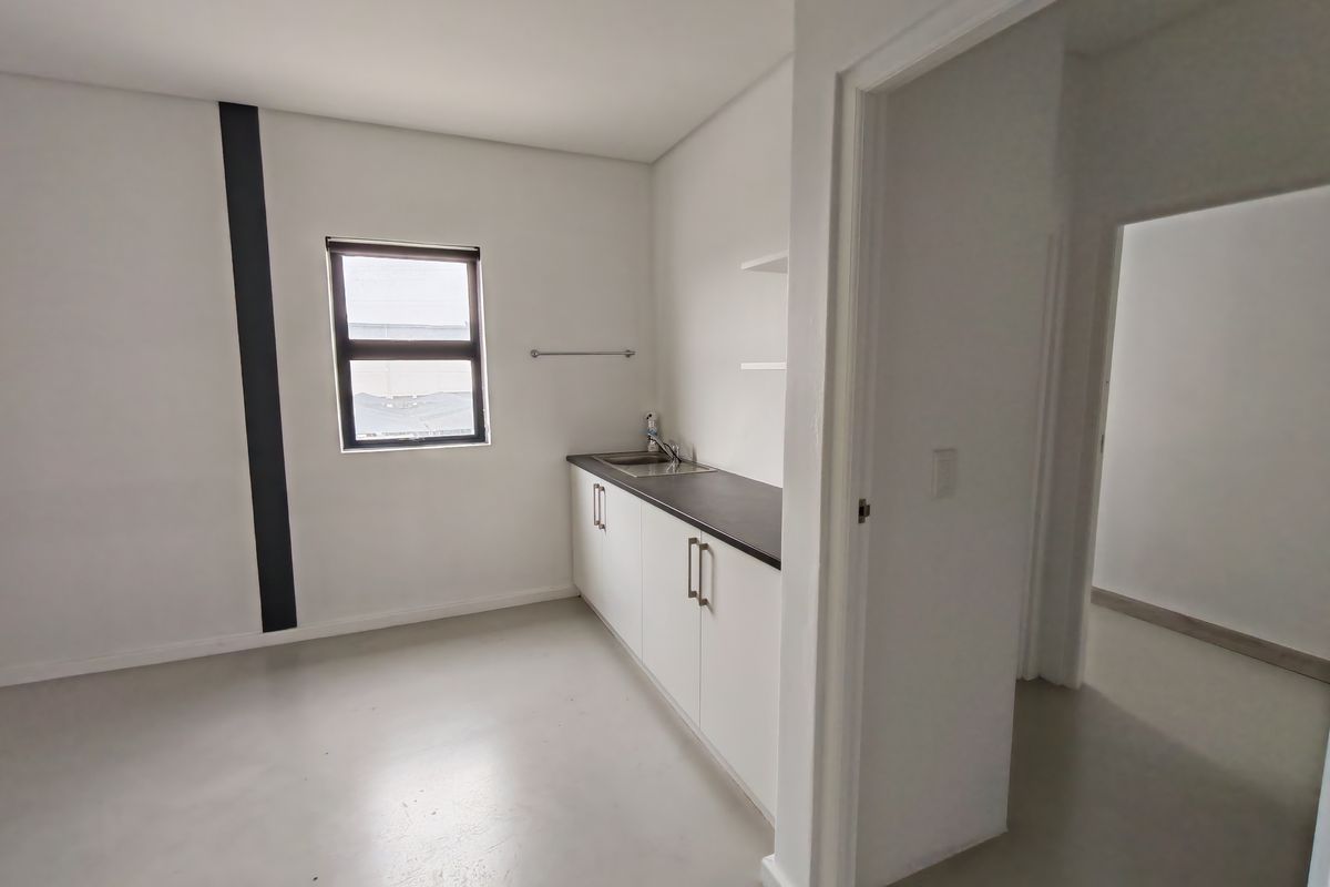 A clean, minimalist kitchenette area featuring white cabinetry, a dark countertop, a stainless steel sink, and polished concrete flooring.