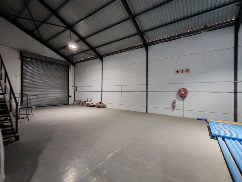 Spacious industrial warehouse unit featuring high ceilings with exposed steel trusses, concrete flooring, and a large roller shutter door for easy access.