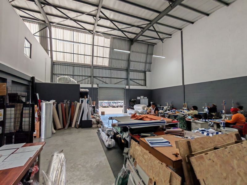 Spacious industrial warehouse unit featuring high ceilings with exposed steel trusses, concrete flooring, and a large roller shutter door for vehicle access.