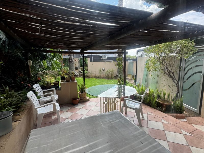 A covered outdoor patio area featuring a rustic thatched pergola, tiled flooring, and a built-in masonry counter, overlooking a small garden space.