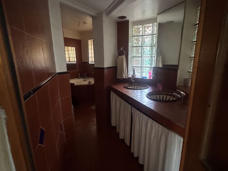A dated bathroom featuring a double vanity with built-in basins, tiled countertops, and a separate bathtub area with glass block windows.