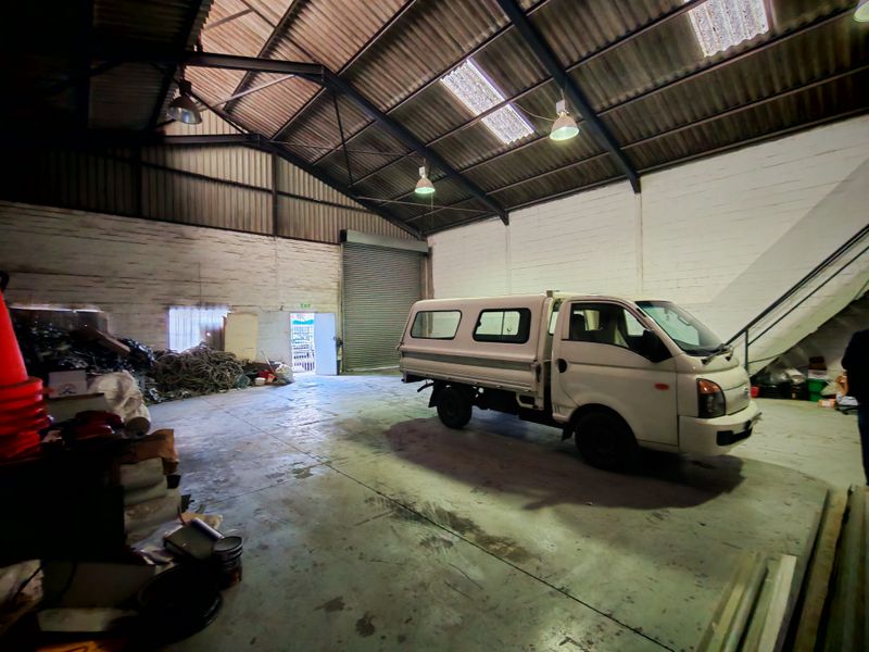 Spacious industrial warehouse unit featuring high ceilings with exposed steel trusses, concrete flooring, roller shutter access, and natural light through skylights.