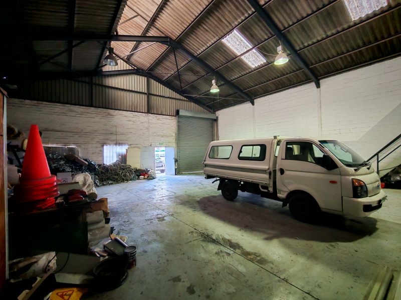 Spacious industrial warehouse or garage featuring high ceilings with exposed steel trusses, concrete flooring, and a large roller shutter door for vehicle access.