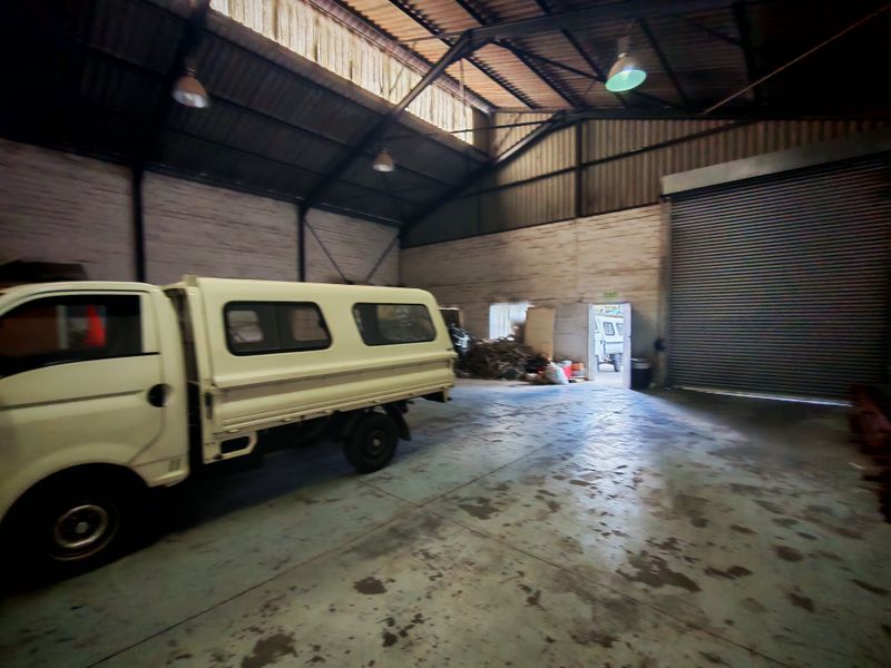 Spacious industrial warehouse or garage featuring high exposed steel roof trusses, concrete flooring, and a large roller shutter door for vehicle access.