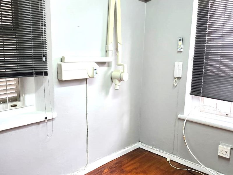 A functional commercial office or clinical space featuring timber flooring, wall-mounted dental imaging equipment, split-system air conditioning, and dual windows with venetian blinds.
