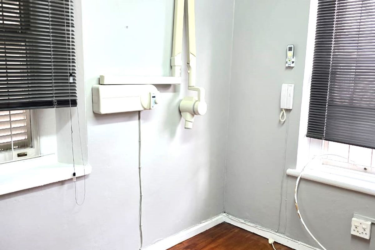 A functional commercial office or clinical space featuring timber flooring, wall-mounted dental imaging equipment, split-system air conditioning, and dual windows with venetian blinds.