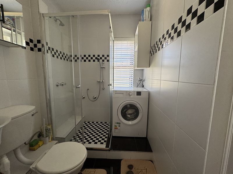 Compact bathroom featuring a glass shower enclosure, toilet, and integrated laundry space with tiled finishes and a checkerboard accent border.