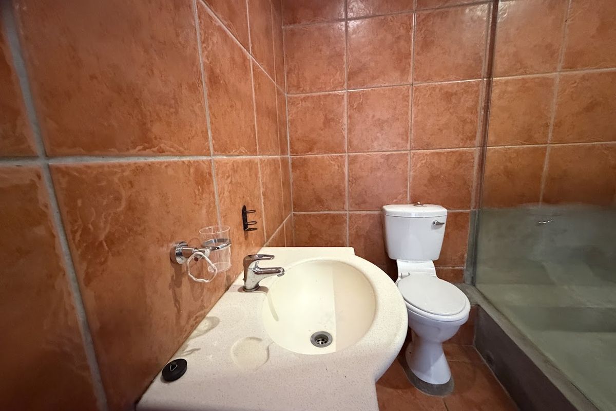 A compact bathroom featuring dated orange ceramic wall and floor tiles, a pedestal washbasin, a standard toilet, and a glass shower screen.