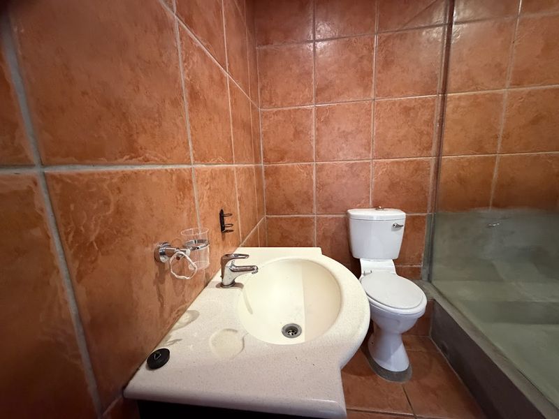 A compact bathroom featuring dated orange ceramic wall and floor tiles, a pedestal washbasin, a standard toilet, and a glass shower screen.