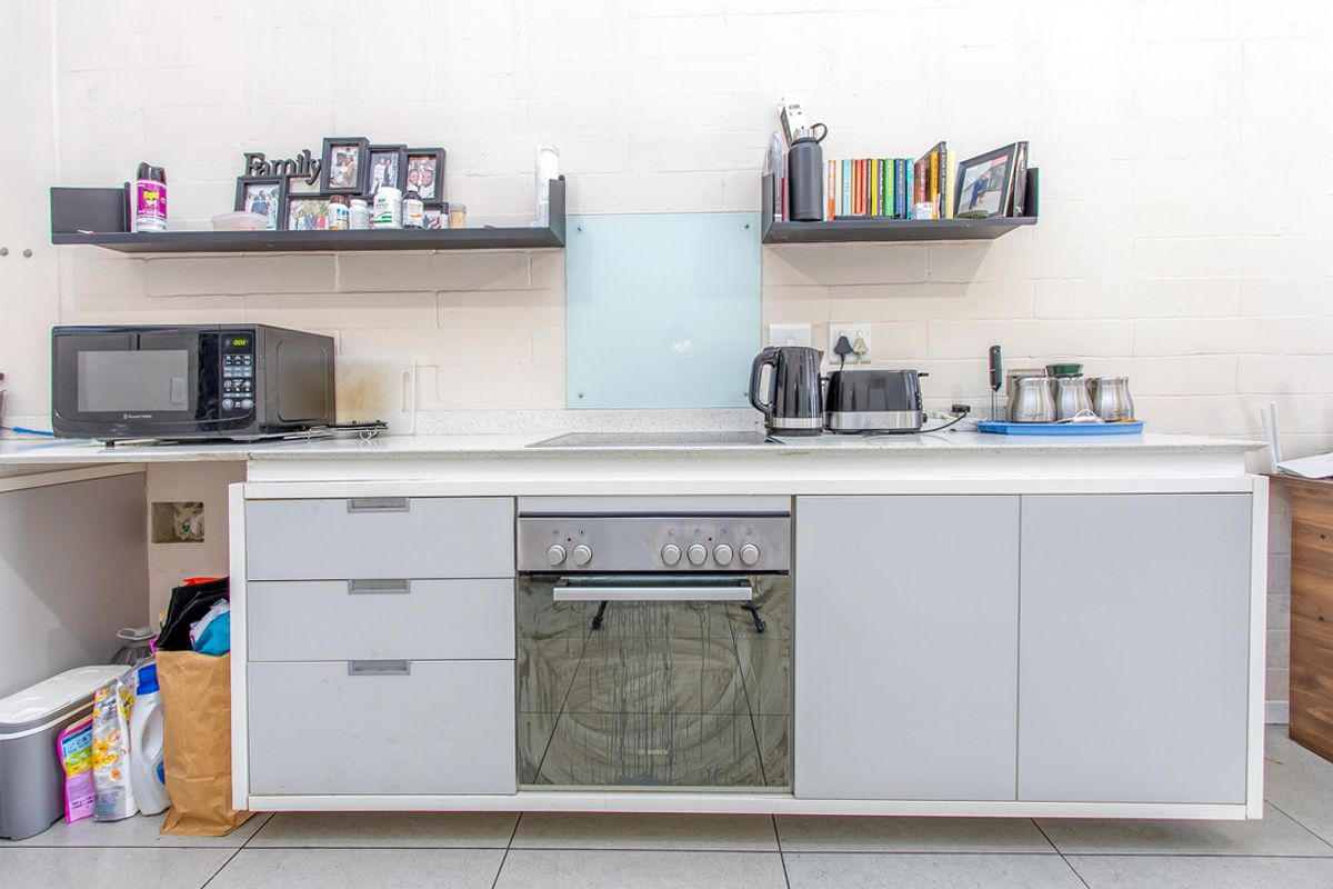A functional kitchen area featuring light grey cabinetry, a built-in oven, electric hob, and stone-look countertops with floating wall shelving.
