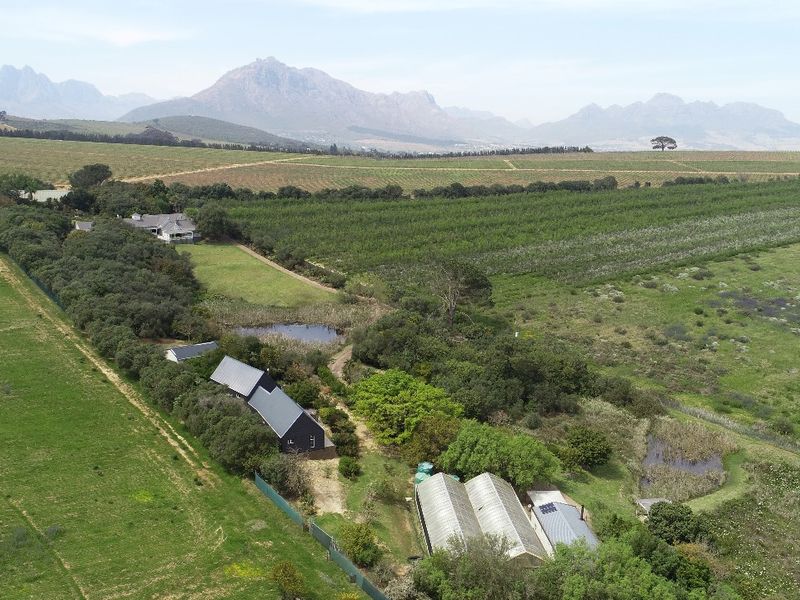 Aerial view of a small holding property featuring agricultural land, multiple farm buildings, greenhouses, and a natural pond set against a mountain backdrop.