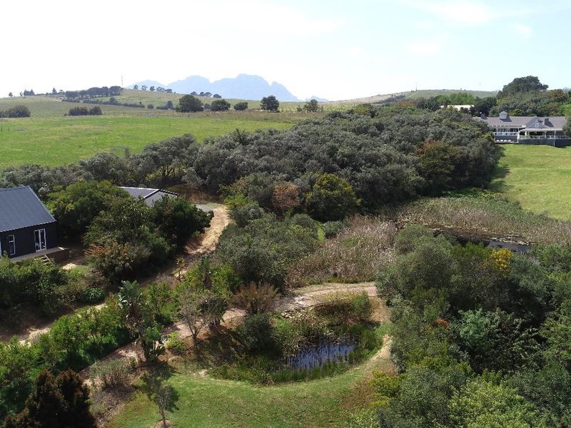 Aerial view of a small holding property featuring a dark-toned dwelling, a natural pond, extensive landscaped grounds, and surrounding rural landscape.