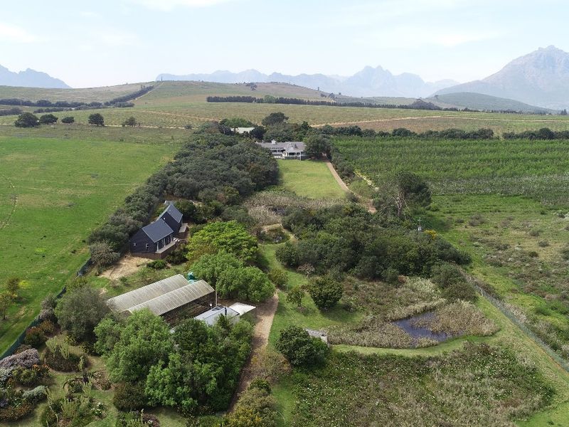 Aerial view of the small holding property featuring multiple structures, a greenhouse, a pond, and expansive surrounding agricultural land with mountain views.